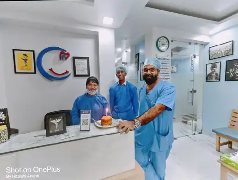 Three clinic staff in blue scrubs standing behind the reception counter, smiling for the camera with the clinic's blue and red logo and framed certificates on the wall behind them.