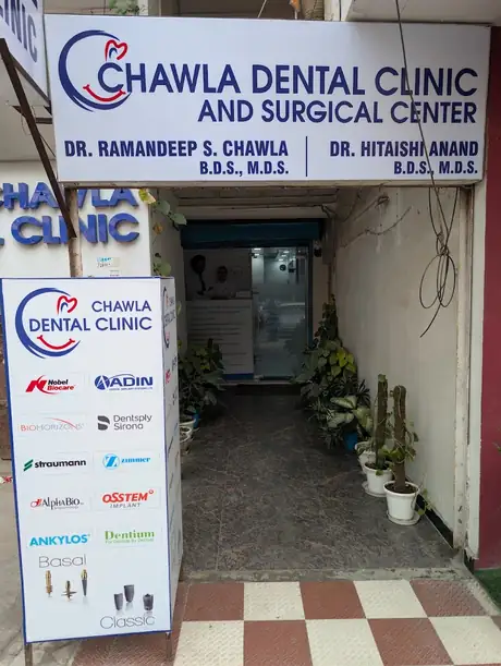 The clinic's white signboard reading Chawla Dental Clinic and Surgical Center above Dr. Ramandeep S. Chawla and Dr. Hitaishi Anand's names, over a tiled entrance walkway lined with potted plants.