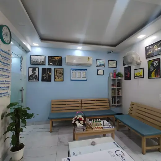 A small blue-walled waiting room with two wooden benches, framed art on the walls, a wall-mounted air conditioner, and a board listing the clinic's dental specialities.